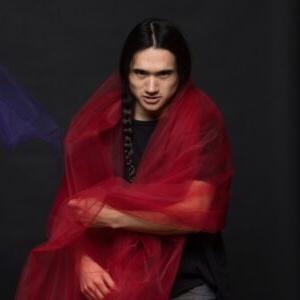 Micah Huang has long black hair in a braid and wraps red tulle fabric around his shoulders while looking intensely at the camera. Huang is wearing a black shirt and jeans and stands against a dark gray background.