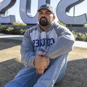 Michael Griggs sits on a grass hill with part of large CSUSB letter signs in the background. Griggs has a short black beard and wears a baseball cap, gray and white sweatshirt, and blue jeans.